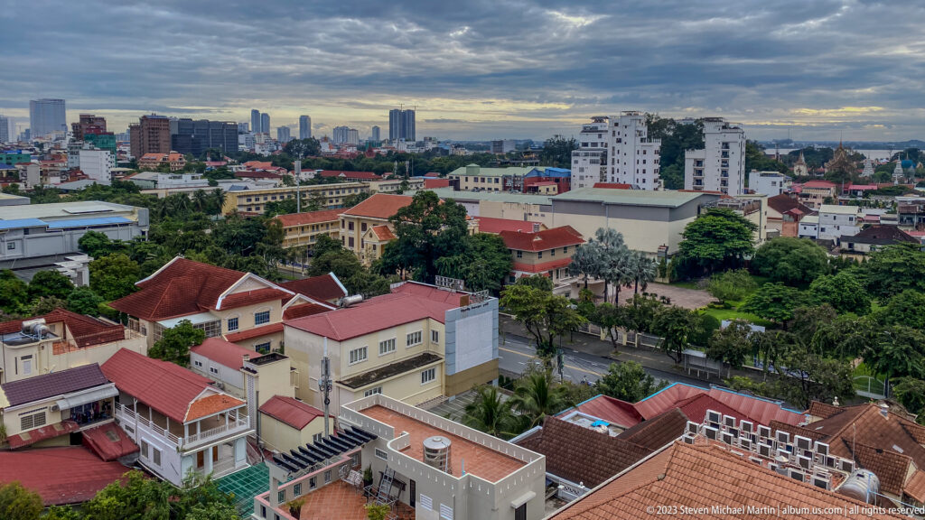 Elevated Phnom Penh view over villas, red roofs, trees, roads, and distant high-rise buildings beneath dark clouds.