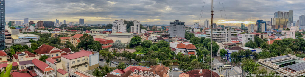 Wide Phnom Penh panorama with red roofs, trees, mid-rises, modern towers, and sunset light beneath heavy clouds.