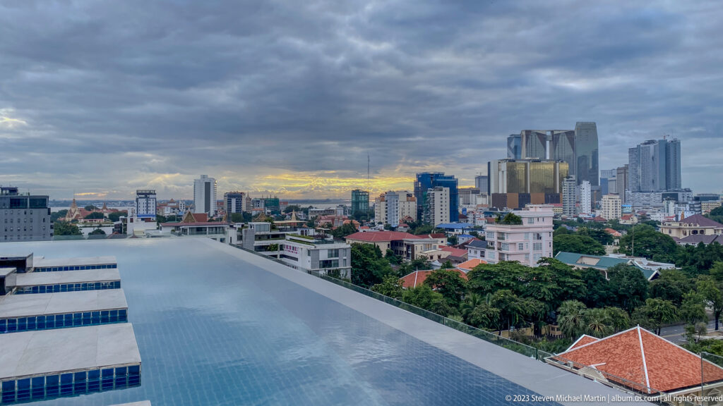 Rooftop infinity pool overlooking red roofs, trees, and Phnom Penh high-rises beneath a cloudy sky.