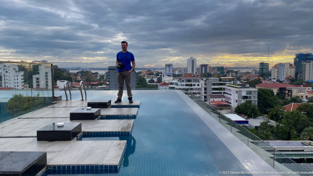 Man standing beside a rooftop infinity pool with Phnom Penh’s skyline and dark evening clouds behind him.