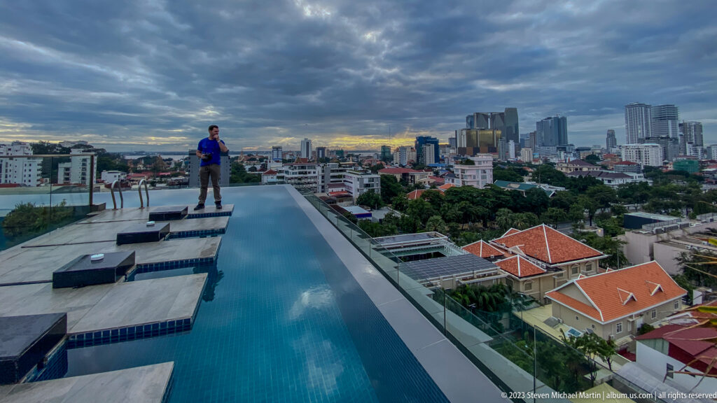 Wide rooftop infinity pool with a distant man at the far end and Phnom Penh skyline beyond.