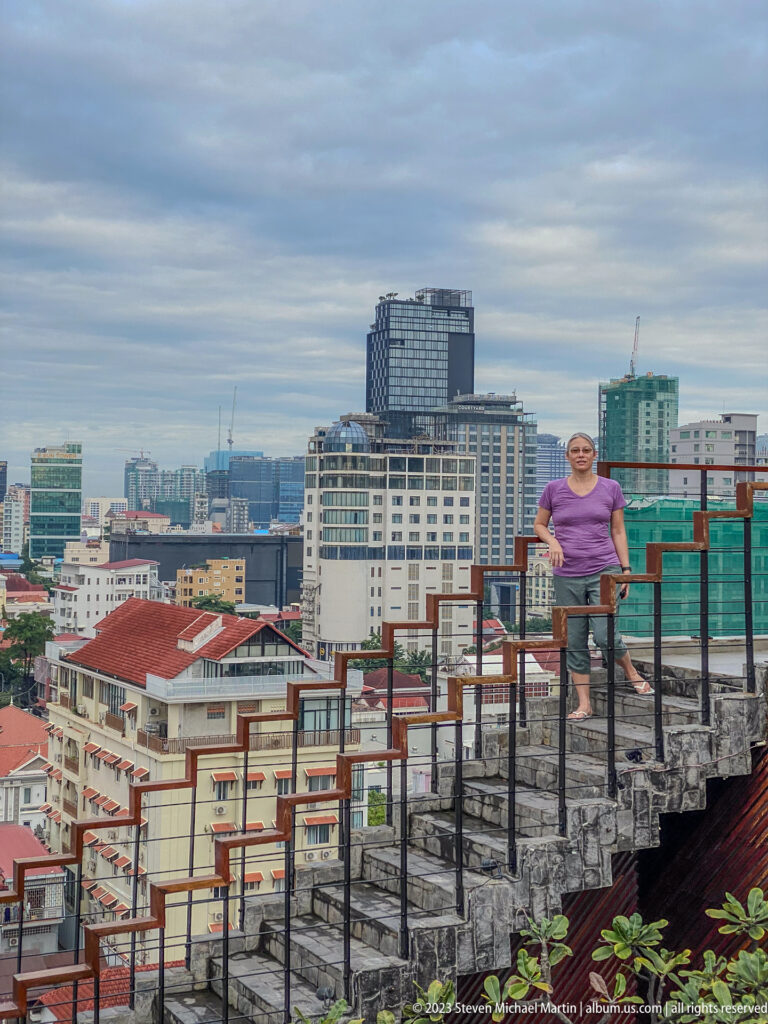 Woman standing among exposed steel bars on an unfinished rooftop with Phnom Penh buildings behind her.