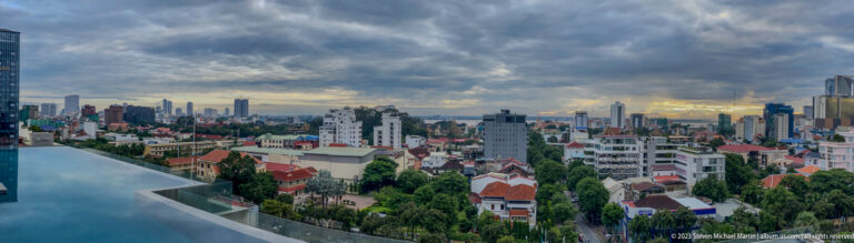 Rooftop pool edge overlooking Phnom Penh’s low-rise districts and distant towers beneath dark storm clouds.