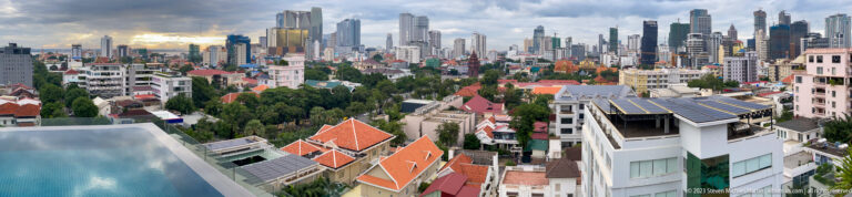 Wide Phnom Penh panorama with rooftop pool, red-roofed compounds, trees, solar panels, and dense high-rises.