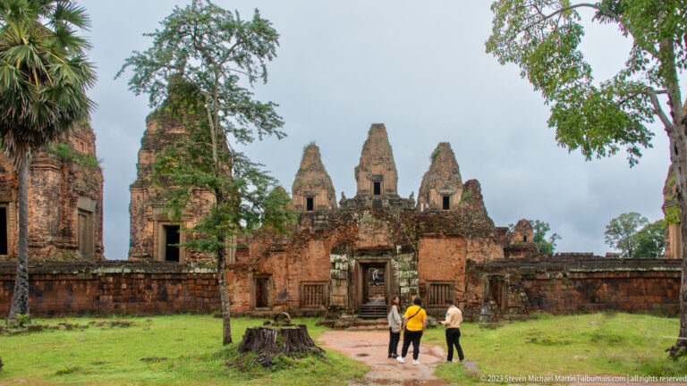 Pre Rup temple entrance with brick sanctuary towers beyond a grassy forecourt and three visitors walking toward the doorway.