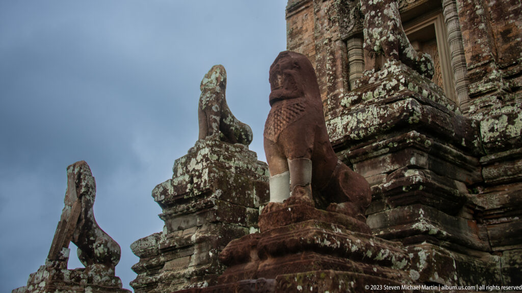 Sandstone lion guardian on a temple terrace beside a weathered brick tower under a dark sky.