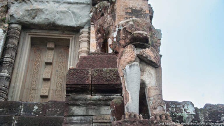 Weathered stone lion guardian beside a pale temple doorway, viewed from below.