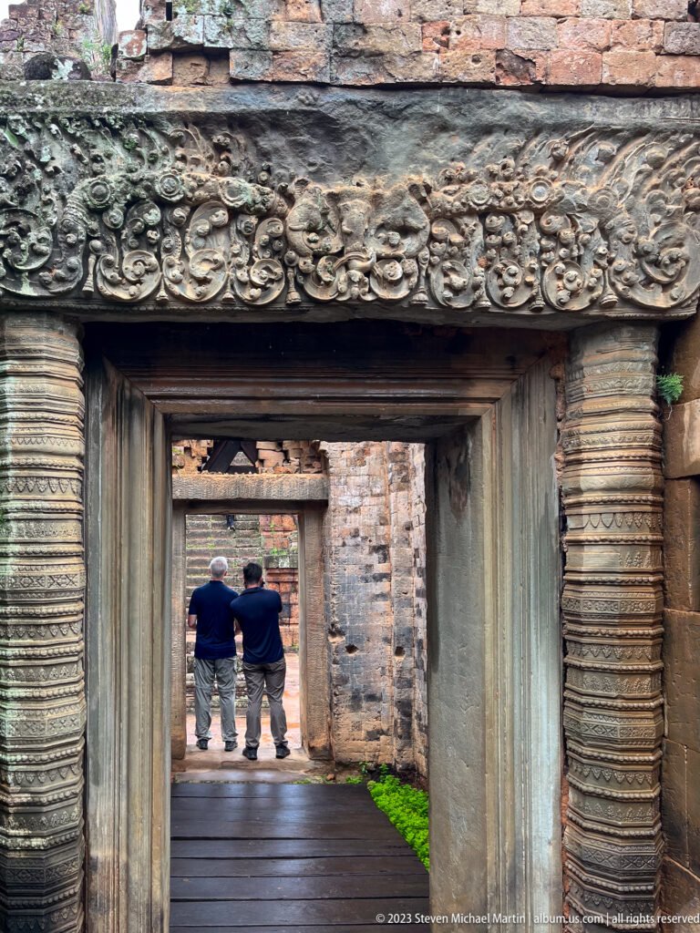 Two visitors framed at the end of aligned carved stone doorways and a wooden walkway.