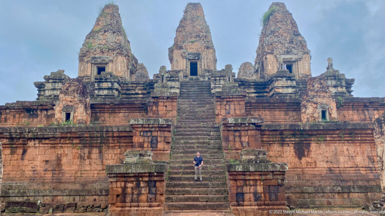 Single visitor climbing the steep central staircase beneath three brick sanctuary towers at Pre Rup.