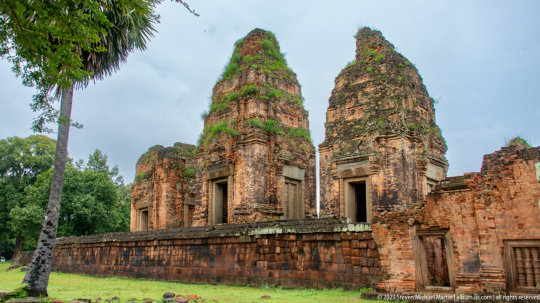 Two tall weathered brick towers at Pre Rup rise behind a low enclosure wall beside a palm tree.
