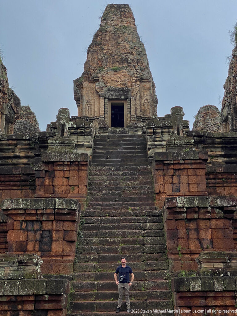 Man seated on the lower steps of a steep temple stair below a brick shrine with a dark doorway.