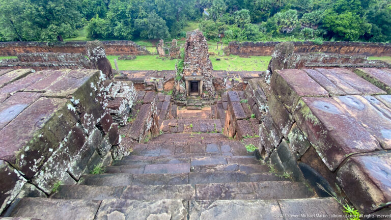 Downward view along steep stone steps toward a small brick shrine, courtyard, and green trees beyond.