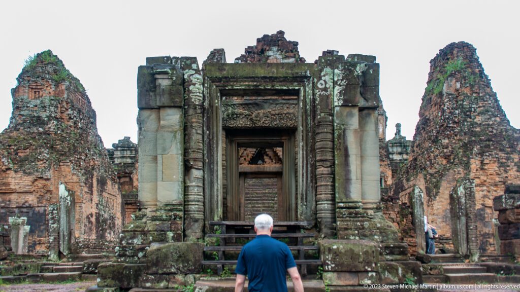 Visitor seen from behind facing a tall carved stone doorway between weathered brick towers at Pre Rup.