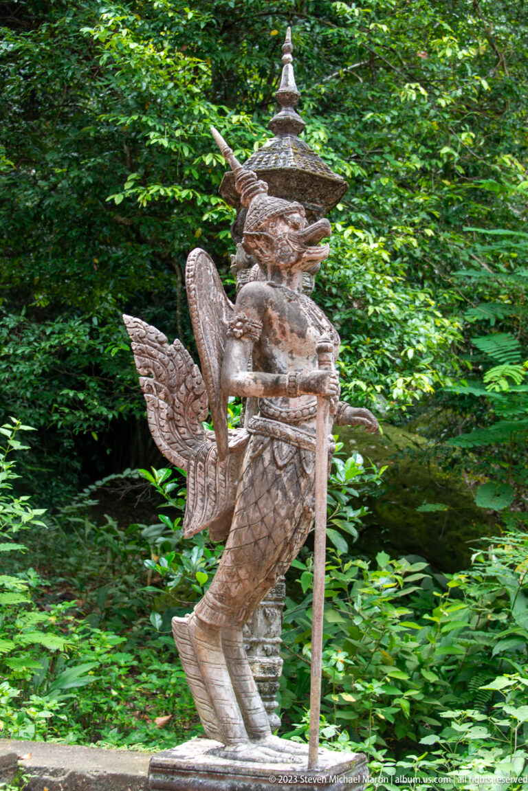 Profile of a crowned winged guardian statue holding a staff in a forest setting.