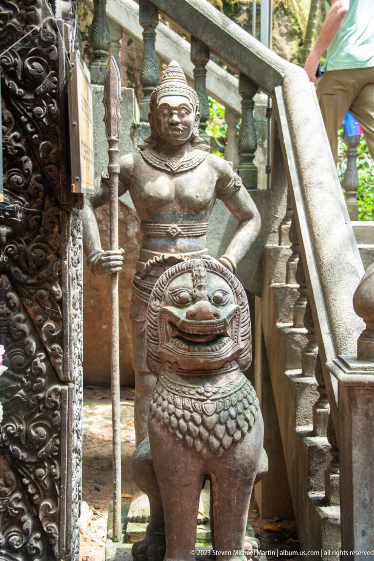 Standing armed guardian behind a seated lion beside carved stairs and doorway.