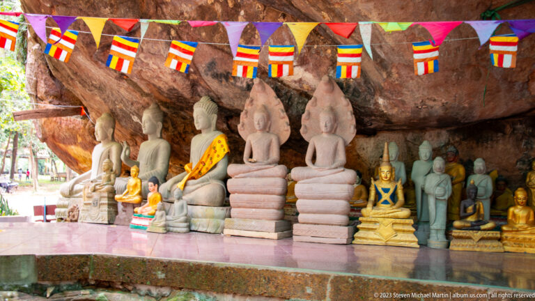 Rows of seated Buddha statues beneath a rock overhang with Buddhist flags.