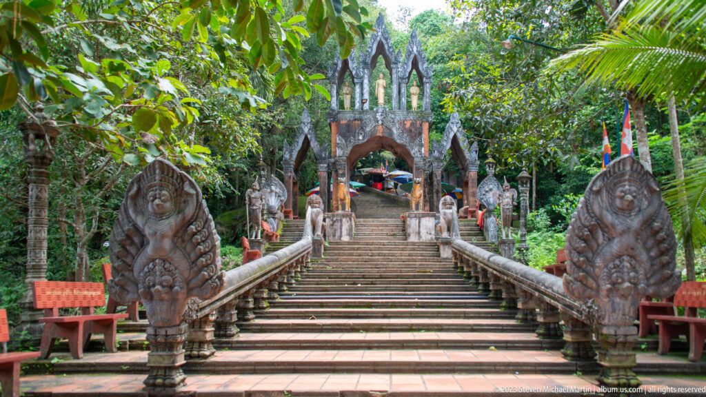 Stone stairway through trees leading to an ornate gateway flanked by naga balustrades.