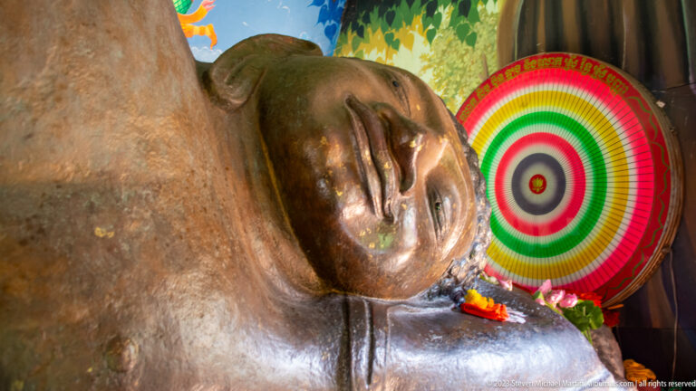 Close view of a reclining Buddha’s face beside a striped ceremonial fan and flowers.
