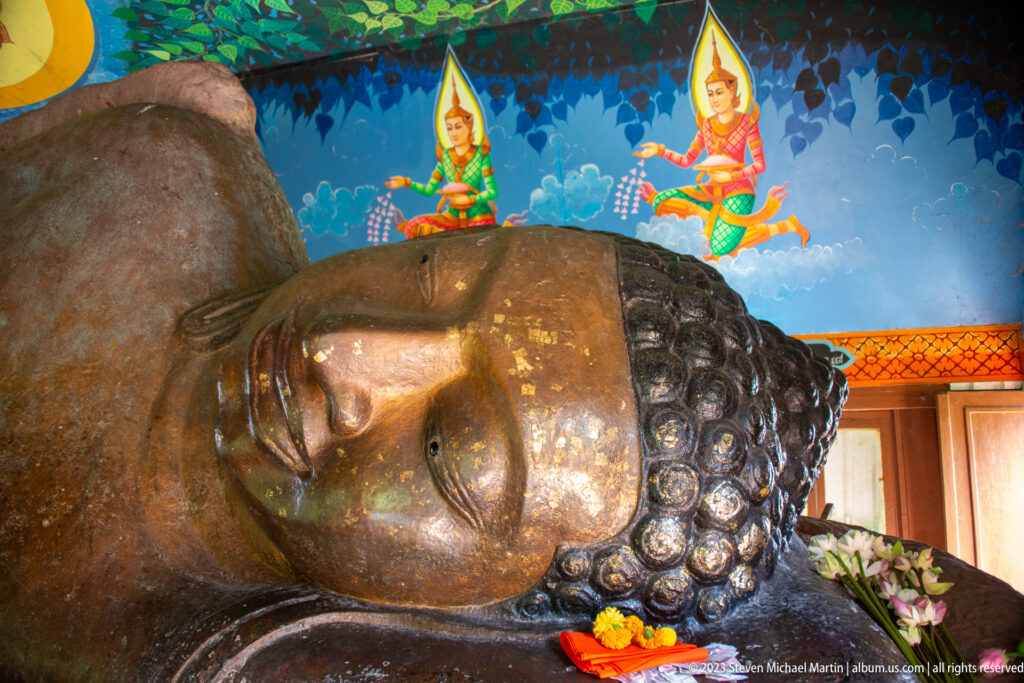 Head and upper torso of a reclining Buddha against a blue mural with celestial figures.