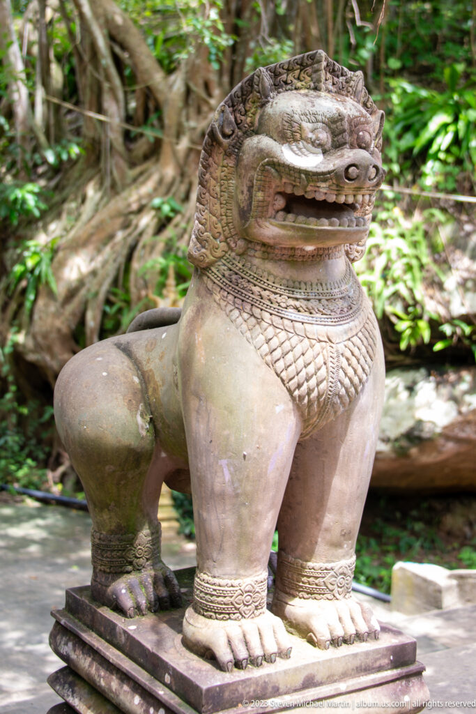 Pale seated guardian lion on a low plinth in a wooded setting.