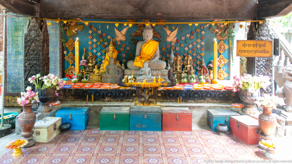 Wide altar with Buddha statues, flowers, candles, and colorful chests.