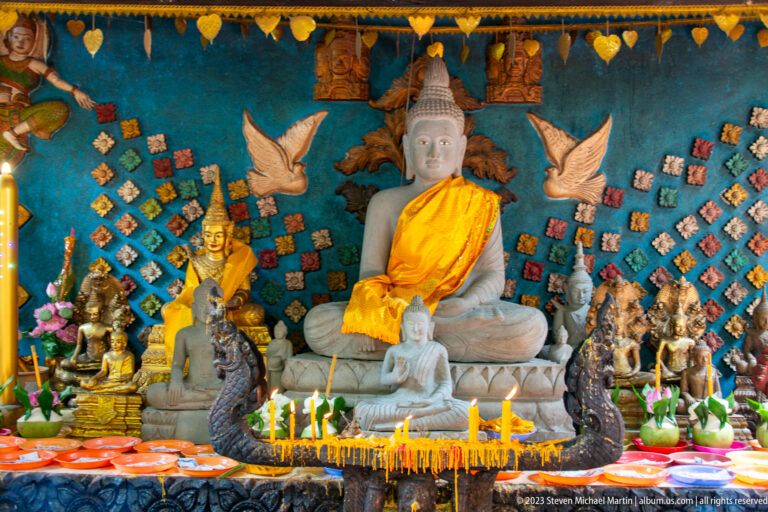 White seated Buddha in a saffron robe surrounded by smaller figures, candles, and flowers.