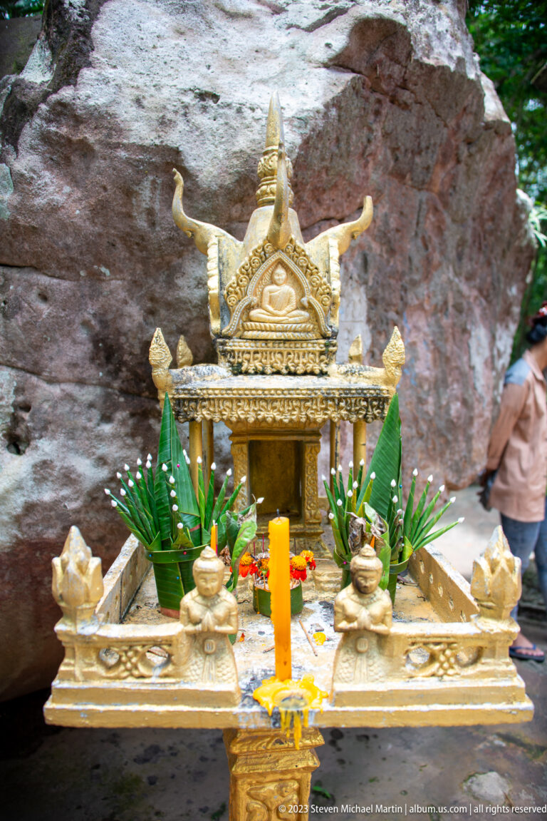 Gilded miniature shrine with candles and leaf offerings set before a large boulder.