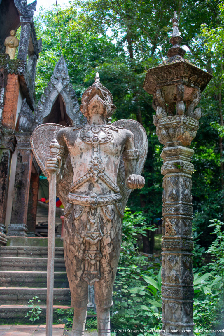 Winged crowned guardian holding a staff beside an ornate carved column.