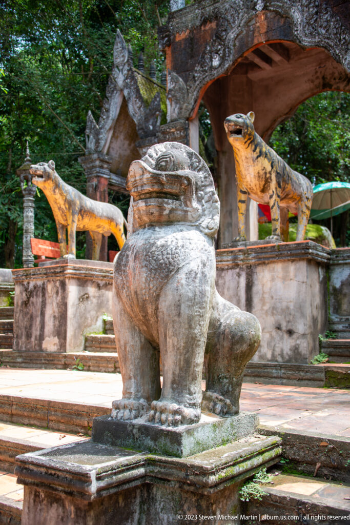 Seated stone guardian lion with painted tiger statues beyond an arched gateway.