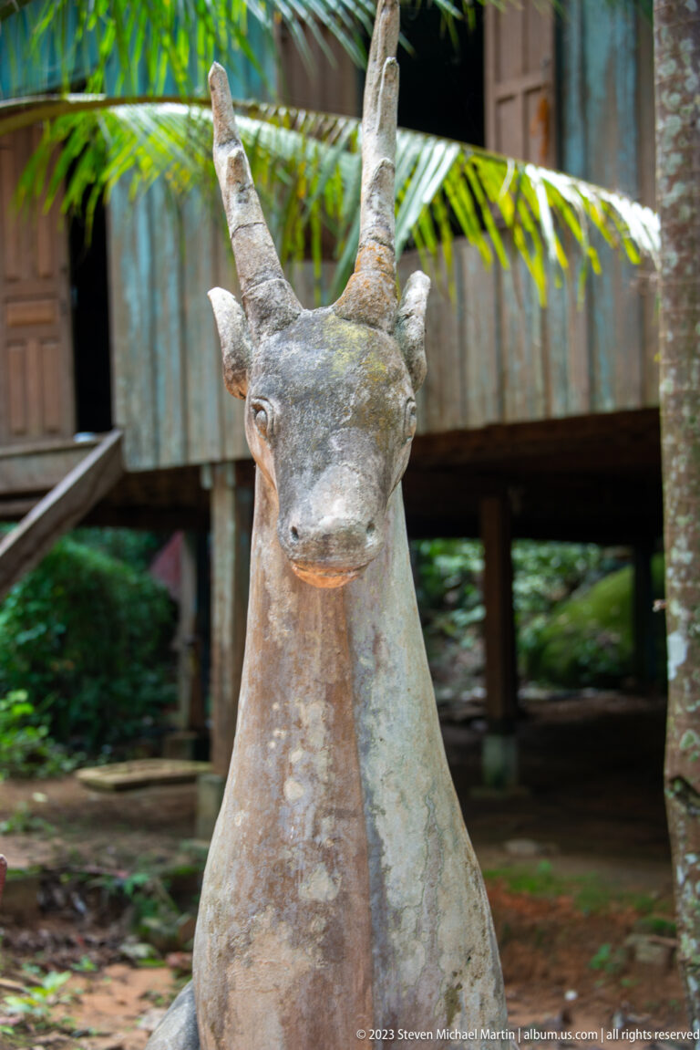 Frontal view of a slender horned animal sculpture before a raised wooden building.