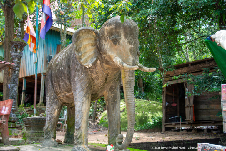 Life-size standing elephant sculpture with tusks and trunk beside monastery buildings.