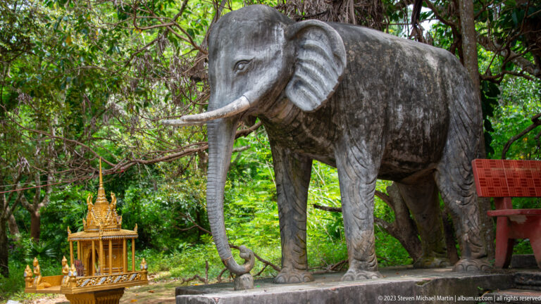 Weathered standing elephant sculpture beside a small gilded shrine in woodland.
