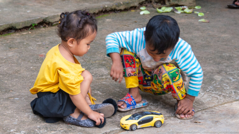 Two young children crouching on pavement and playing with a yellow toy car.