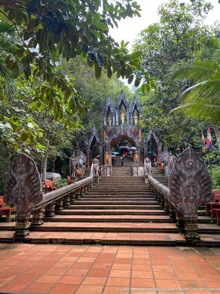 Broad symmetrical stairway with naga finials leading uphill to an ornate gateway.