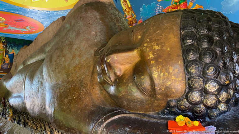 Close angled view of the reclining Buddha’s face, curled hair, shoulder, and marigold offerings.