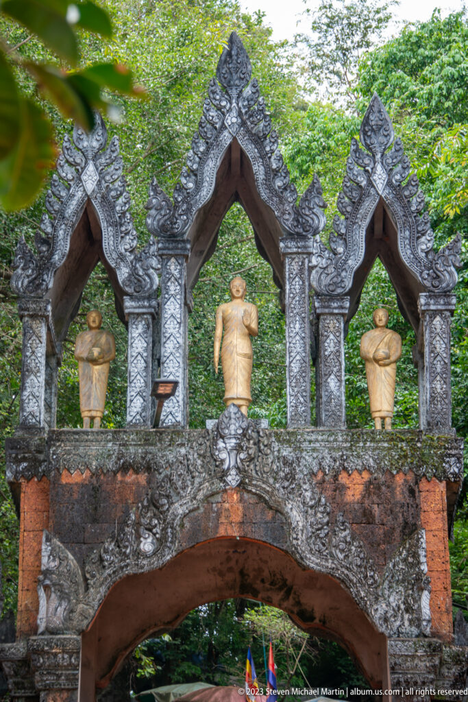 Dark ornamental gateway with three pointed gables and standing Buddha figures above an arch.