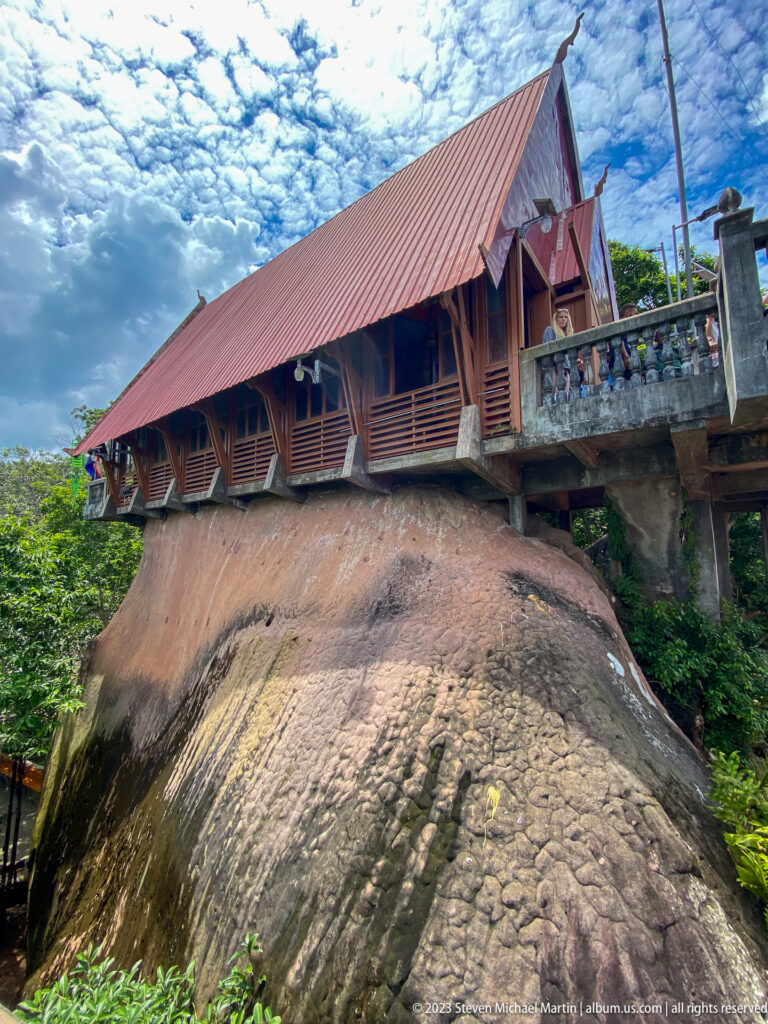 Wooden temple hall with a steep red roof constructed above a massive rock outcrop.