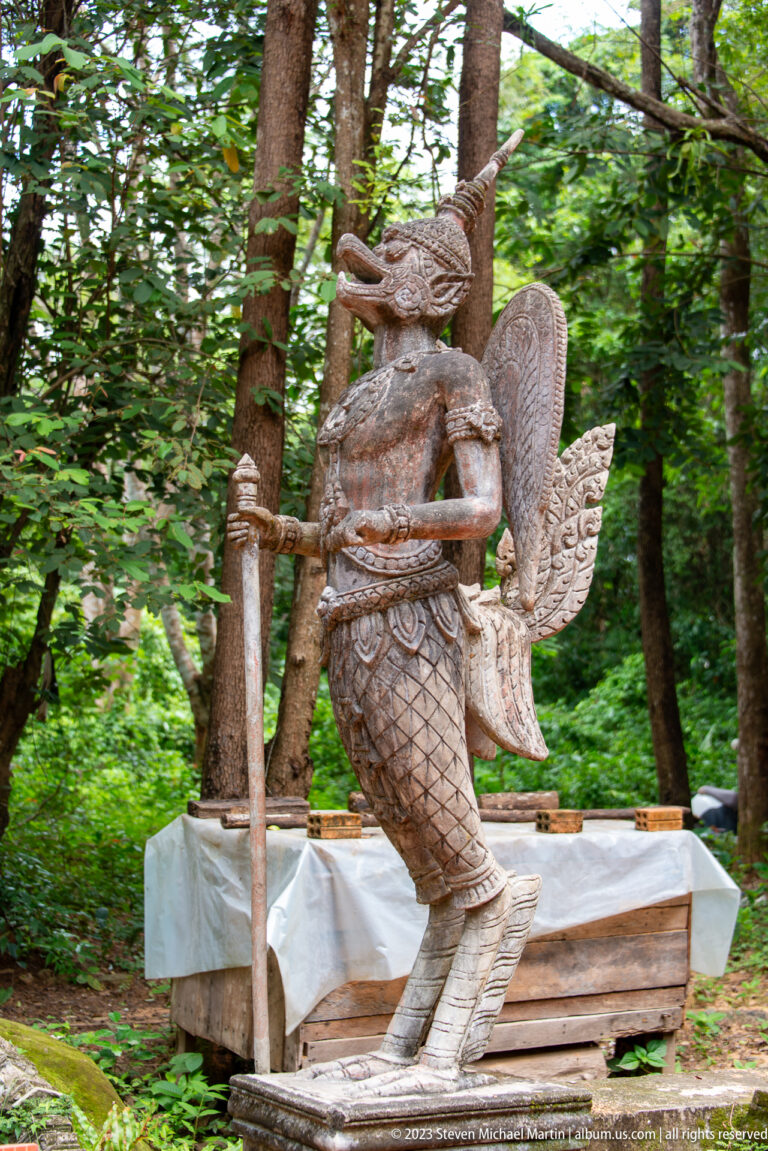 Weathered bird-bodied guardian statue holding a long staff beneath trees.