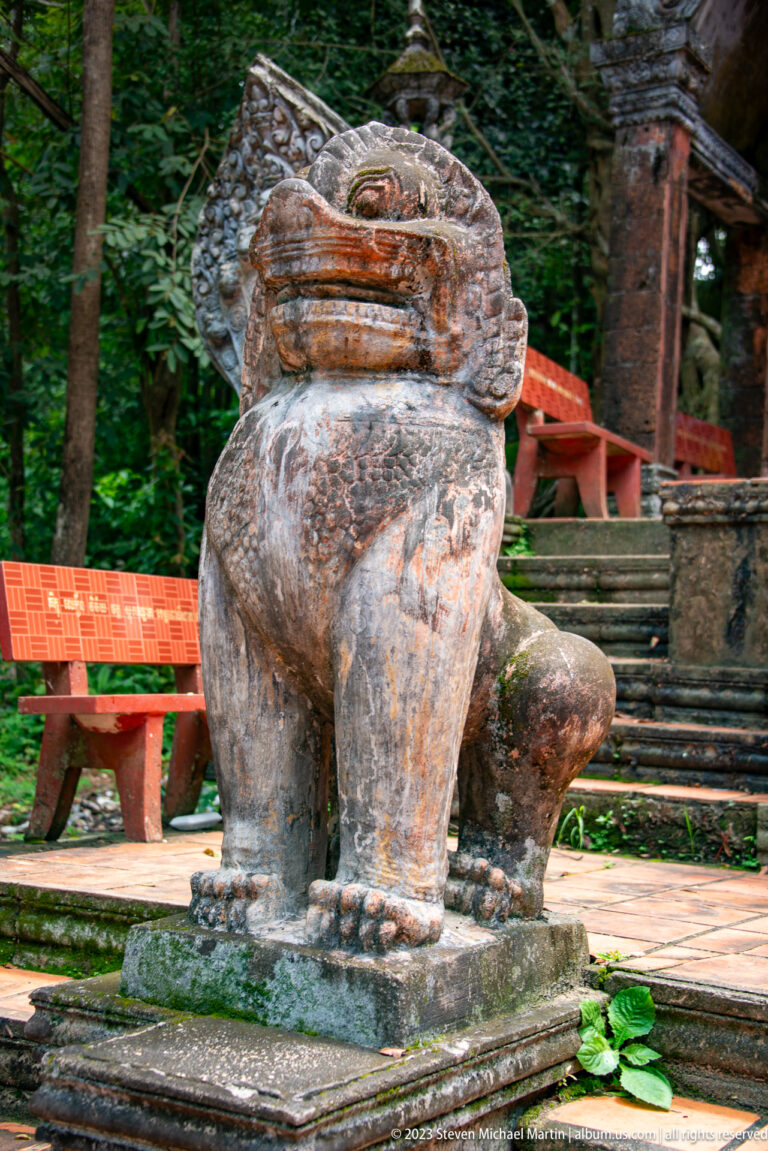 Weathered seated stone lion on a square base near temple steps.
