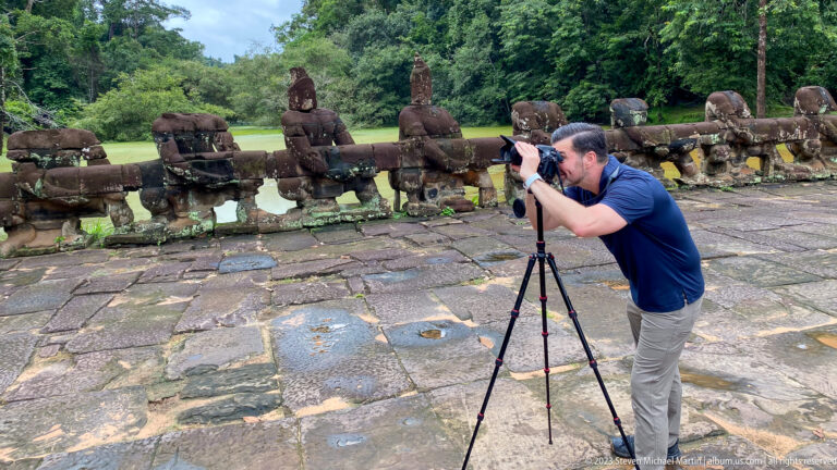 Male visitor using a tripod-mounted camera beside carved stone guardians along the temple causeway.