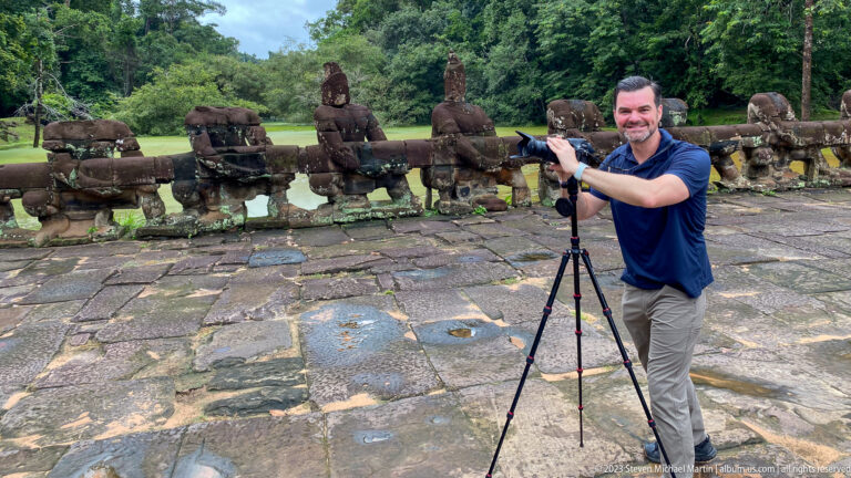 Smiling male visitor beside a camera tripod with rows of stone guardians behind him.