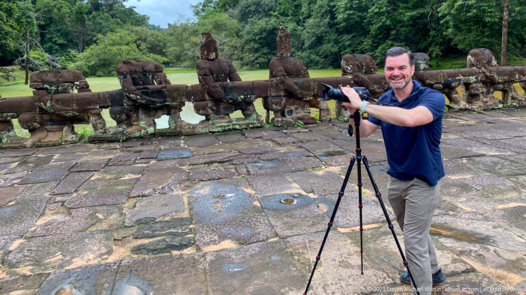 Male visitor gesturing beside a camera tripod with carved causeway guardians behind.