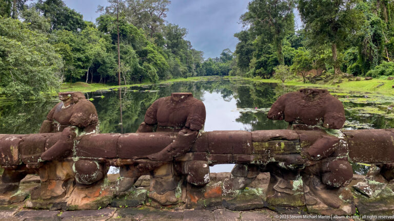 Rear view of three damaged stone guardians facing a calm forested moat.