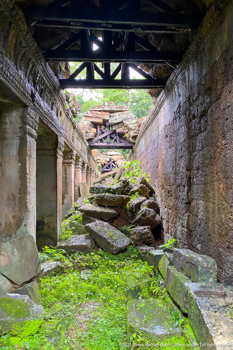 Long ruined corridor with fallen sandstone blocks, bright green moss, and partial roof supports overhead.