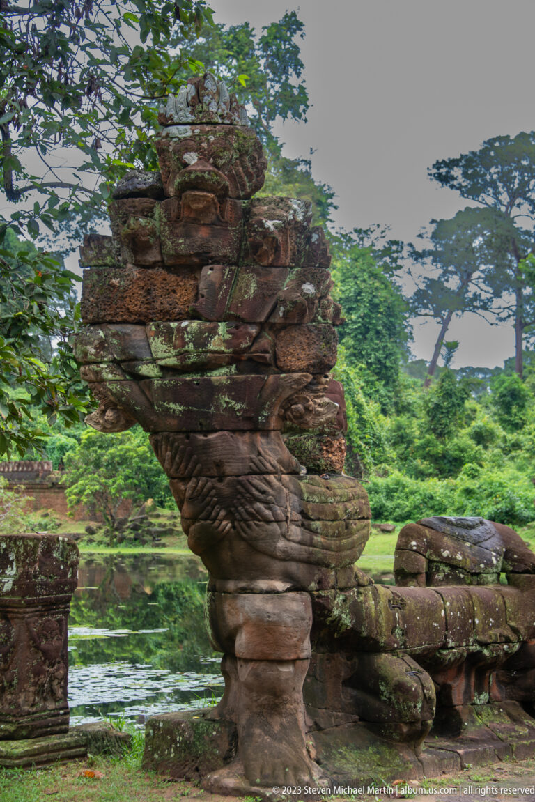 Large weathered sandstone guardian with avian features beside a row of smaller causeway sculptures.