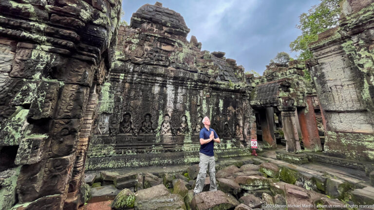 Male visitor standing on fallen blocks before a richly carved sandstone courtyard wall.