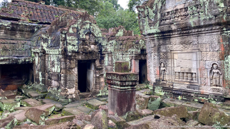 Ruined sandstone courtyard with carved doorways, blind windows, female figures, and fallen blocks.