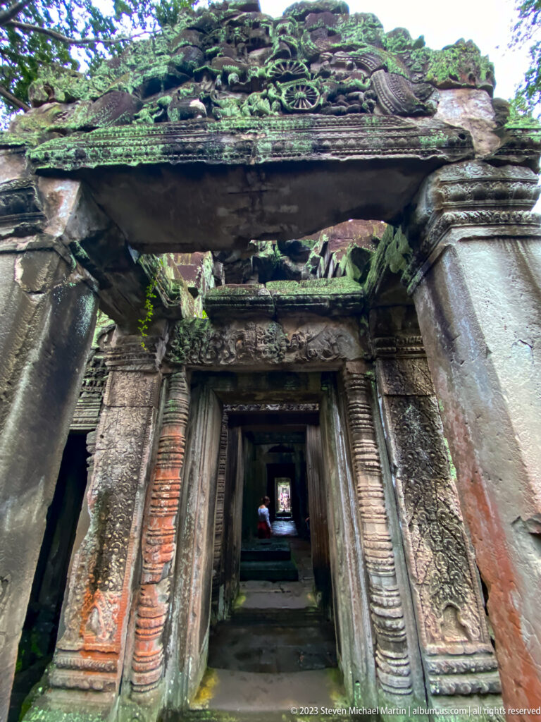 Damaged carved sandstone doorway with collapsed pediment blocks and aligned chambers beyond.