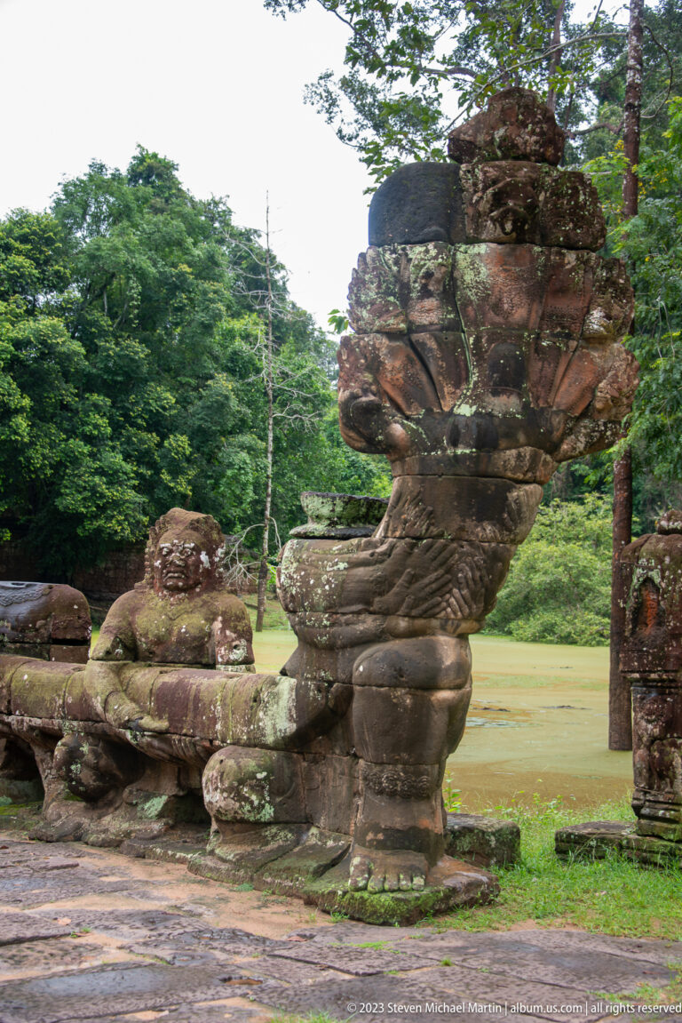 Three-quarter view of a massive avian guardian sculpture beside the temple causeway and pond.