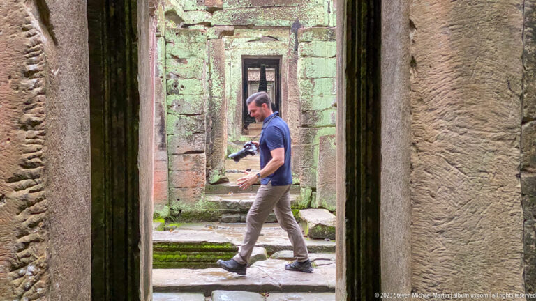 Male visitor carrying a camera while walking through aligned sandstone temple doorways.